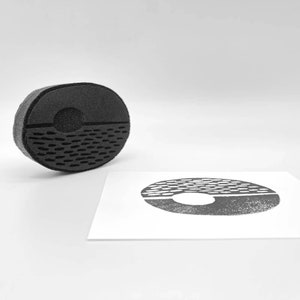 Può includere: Un timbro nero di forma ovale con un design geometrico. Il timbro è diviso in due sezioni, con un'indentazione circolare nella metà superiore e un motivo strutturato nella metà inferiore. Un'impressione del timbro è su una carta bianca.
