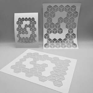 May include: Three white paper stencils with a geometric cube design. The stencils are in different sizes and shapes, with the largest one flat on the surface and the other two slightly curved. The design is cut out of the paper.