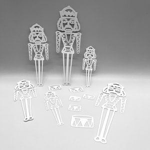 May include: White, die-cut paper Christmas nutcracker figures in various sizes, along with drum and banner shapes. The figures have detailed facial features, uniforms, and crowns. The objects are arranged on a light gray background.
