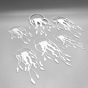 May include: Six white paper cutouts of jellyfish are arranged on a grey background. Each jellyfish has a bell-shaped body with intricate details and long, flowing tentacles. The cutouts are in various orientations, creating a dynamic composition.