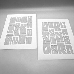 May include: Two white stencils with a geometric design. Each stencil features a grid of rectangular shapes with curved lines and oval shapes cut out. The stencils are on a gray background.