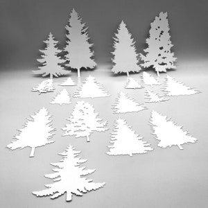 May include: A collection of white paper cut-out evergreen trees of various sizes. The trees are arranged on a light gray surface, with some standing upright and others lying flat. The cut-outs have detailed branch and needle patterns.