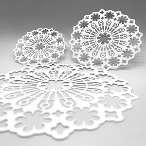 May include: Three white, laser-cut paper doilies with intricate, symmetrical designs. The doilies vary in size, with a large one in the foreground and two smaller ones behind it. The designs feature floral and geometric patterns, creating a delicate and ornamental aesthetic.