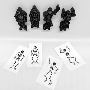 Könnte beinhalten: Schwarze, skelettförmige Dekorationen und temporäre Tattoos. Die Dekorationen sind dreidimensional und haben ein mattes Finish. Die Tattoos sind auf weißen rechteckigen Blättern gedruckt. Die Skelette sind in verschiedenen Posen, ideal für Halloween.
