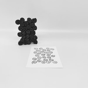 Puede incluir: Una escultura abstracta negra con forma de nube, con numerosos diseños en espiral. Una tarjeta blanca con un patrón de espirales negras está frente a la escultura. El fondo es blanco liso.
