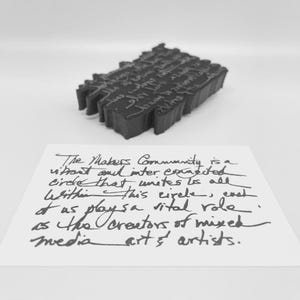 Puede incluir: Imagen en blanco y negro de una escultura oscura y texturizada con texto grabado, colocada sobre una tarjeta blanca. La tarjeta muestra texto manuscrito: "The Makers Community is a vibrant and interconnected circle..." Tema: arte y comunidad.