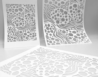 Op de afbeelding: Drie witte papieren uitsparingen met een gelaagd ontwerp met organische vormen en vloeiende lijnen. De uitsparingen variëren in grootte en zijn zo gerangschikt dat de ingewikkelde details van het ontwerp te zien zijn. De patronen omvatten cirkels en golvende lijnen.