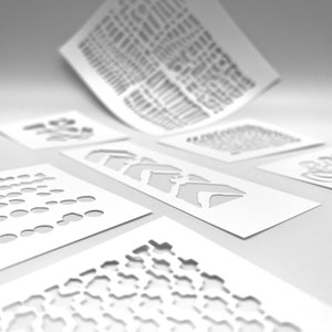 Op de afbeelding: Een verzameling witte papieren sjablonen met diverse uitgesneden ontwerpen, waaronder geometrische vormen en patronen. De sjablonen zijn gerangschikt op een wit oppervlak, met één iets opgetild.