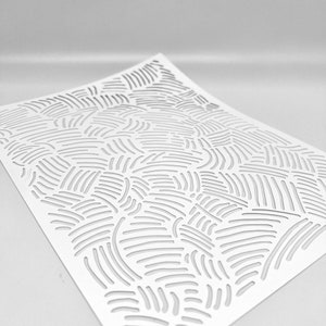 May include: A white paper sheet with a cut-out pattern of curved, parallel lines. The lines are evenly spaced and create a textured, abstract design. The paper is angled, showing the cut-out pattern in detail.
