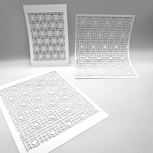 May include: Three white paper stencils with a repeating geometric pattern of squares, circles, and lines. The stencils are arranged at different angles, with one slightly curved. The design is cut out of the paper.
