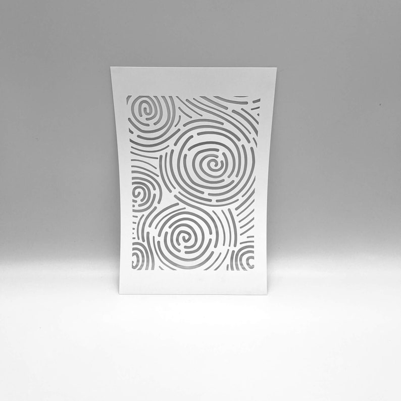Op de afbeelding: Een witte papieren sjabloon met een uitgesneden ontwerp van concentrische cirkels en gebogen lijnen. Het ontwerp is gecentreerd op het papier, met een grijze achtergrond. De sjabloon wordt waarschijnlijk gebruikt voor knutselen of decoratieve doeleinden.
