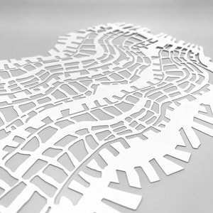 May include: A white paper cut-out of a city map, showing detailed street layouts and building shapes. The layered design gives a 3D appearance. The artwork is set against a light grey backdrop, with shadows adding depth to the design. The overall effect is of a detailed urban landscape.