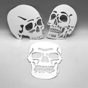 Puede incluir: Tres recortes de calaveras blancas en diferentes posiciones. Dos calaveras están de pie, una frente a la otra, y la tercera está plana. Los recortes presentan estructuras faciales detalladas, incluyendo cuencas de los ojos, cavidades nasales y dientes, creando una estética espeluznante.