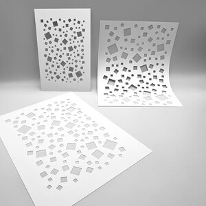 May include: Three white paper cutouts with a pattern of varying sized squares and diamond shapes. The cutouts are arranged at different angles, with one standing upright, one curved, and one lying flat. The background is a light gray.