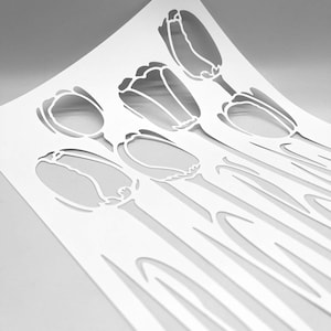 May include: A white paper stencil with a tulip flower design. The stencil has six tulip outlines cut out, with stems and leaves visible. The paper is slightly curved, creating shadows that highlight the design.