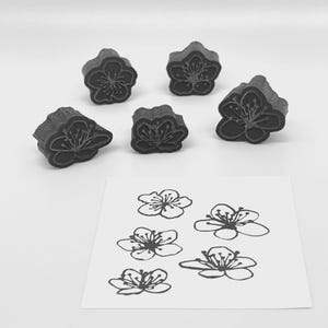 May include: Six dark gray flower-shaped stamps with detailed floral designs are arranged around a white paper with matching flower outlines. The stamps are three-dimensional, while the outlines are flat.