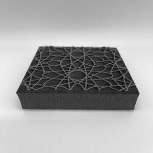 Puede incluir: Un sello de espuma negro y cuadrado con un diseño geométrico de líneas y círculos interconectados. El diseño cubre la superficie superior. El sello se ve desde un ángulo lateral sobre un fondo blanco. El patrón crea un diseño complejo.