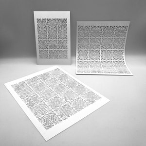 May include: Three white paper cutouts with a repeating geometric pattern. The cutouts are arranged at different angles, with one flat on the surface, and two standing upright. The pattern consists of interlocking circular and square shapes.