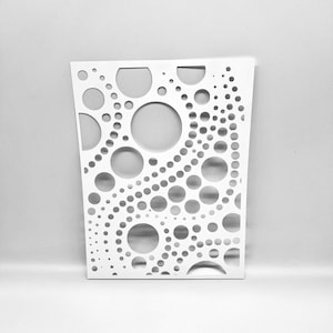 May include: A white, rectangular decorative panel featuring a pattern of circular cutouts of various sizes. The panel has a slightly trapezoidal shape, wider at the top than the bottom. The cutouts vary from small dots to large circles.