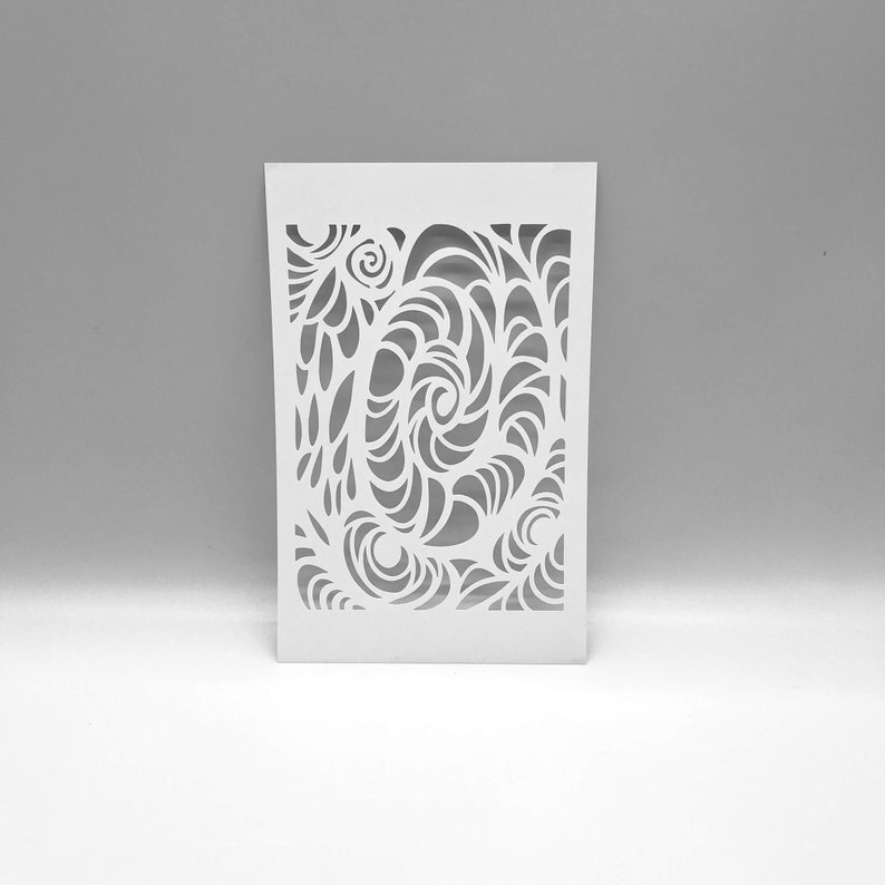 May include: A white stencil with a cut-out design of swirling, organic shapes. The rectangular stencil features a central spiral and flowing, leaf-like patterns. The design is symmetrical and detailed, creating a visually interesting pattern for crafts and art projects.