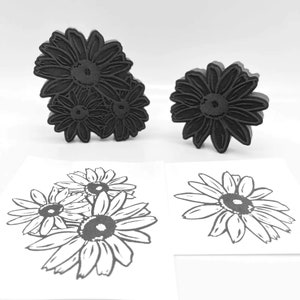 Puede incluir: Dos sellos de goma negra de flores de margarita. El sello más grande tiene tres margaritas, mientras que el sello más pequeño tiene una margarita. Los sellos se muestran junto a dos imágenes impresas de las margaritas.