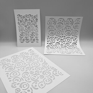 Peut inclure: Trois pochoirs blancs avec un motif de roses en spirale répétitif. Les pochoirs sont disposés à différents angles, dont un à plat et deux debout. Le motif découpé est visible sur un fond neutre.
