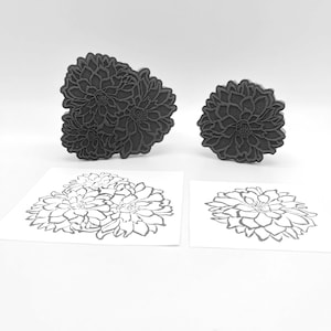Puede incluir: Dos sellos de goma negros con diseños florales. El sello más grande presenta tres flores, mientras que el sello más pequeño tiene una flor. Ambos sellos están sobre una superficie blanca.