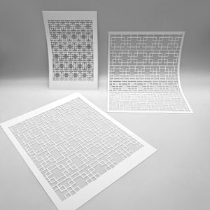 May include: Three white paper stencils with a geometric square pattern. The stencils are arranged at different angles, with one flat on the surface and two standing upright. The design is a repeating pattern of squares and rectangles.