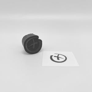 May include: A black rubber stamp with a plus sign inside a circle is displayed next to a small white card with a matching symbol. The stamp has a textured surface and is set against a plain white background.