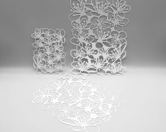 May include: Three white paper cutouts in the shape of cherry blossoms. The cutouts are in different sizes and shapes, with one rectangular, one square, and one circular arrangement of the flowers. The blossoms have detailed petals and stamens.
