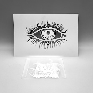 May include: A white stencil depicting a detailed eye with long eyelashes. A small clear bag containing several white eyelash extensions is below the stencil. The image is set against a neutral grey background.