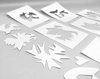 May include: White stencils with various cut-out designs, including abstract shapes, a bird, and the number 15. The stencils are arranged on a light gray surface, providing a clean and organized presentation. Ideal for crafting and artistic projects.