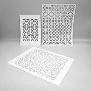 Puede incluir: Tres recortes de papel blanco con patrones florales. Los recortes tienen diferentes formas y tamaños, incluyendo uno rectangular, uno cuadrado y uno curvo. Los recortes están dispuestos sobre una superficie gris claro.