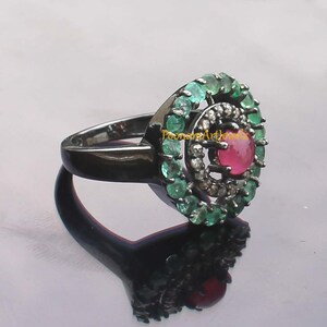 May include: A black metal ring featuring a central red gemstone surrounded by a halo of small white stones. A second halo of green gemstones encircles the center. The ring is displayed on a reflective surface.