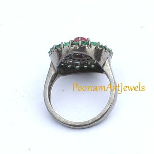 May include: A silver ring featuring a central oval red gemstone, surrounded by a halo of small green gemstones. The ring has a detailed design and is set against a white background. The text "PoonamArtJewels" is visible.