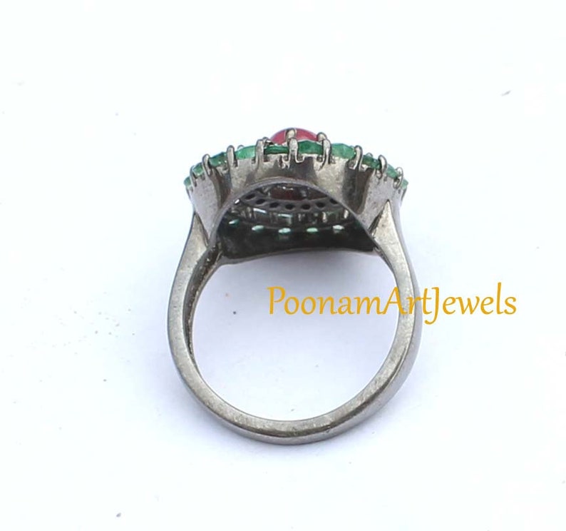 May include: A silver ring featuring a central oval red gemstone, surrounded by a halo of small green gemstones. The ring has a detailed design and is set against a white background. The text "PoonamArtJewels" is visible.
