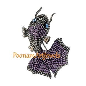 May include: A silver fish brooch set with purple and clear gemstones. The fish has a long tail and a small head with two blue eyes.