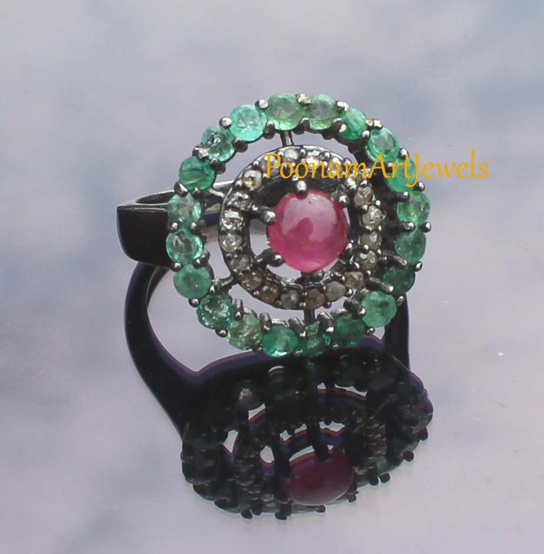 May include: A silver ring featuring a central, round, pink gemstone surrounded by a halo of small diamonds. A second halo of green gemstones encircles the diamonds. The ring's band is black, and the ring is displayed on a reflective surface.