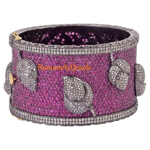 Pode incluir: Um bracelete de prata com acabamento oxidado preto, incrustado com pedras preciosas rosa e brancas. O bracelete apresenta um design floral com folhas e um motivo central. O texto "Poomam Art Jewels" é visível no bracelete.