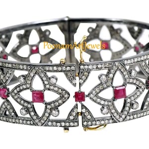 Puede incluir: Un brazalete de plata oxidada negra con un diseño geométrico con detalles de rubí rojo y diamantes blancos. El brazalete tiene bisagras y cierre de broche. "Poonam Art Jewels" está impreso en el brazalete.