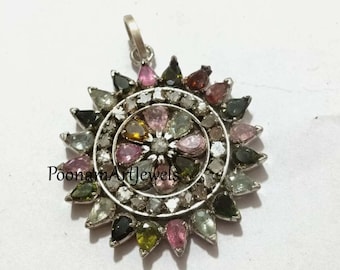 May include: A silver pendant with a circular design featuring a central diamond surrounded by a ring of smaller diamonds and a ring of colorful gemstones. The gemstones are in shades of pink, green, and black.