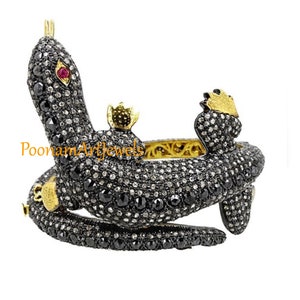Op de afbeelding: Een zilveren en gouden slangenarmband met zwarte en witte diamanten. De slang is opgerold en heeft een kroon op zijn kop. De armband is versierd met een rode robijn.
