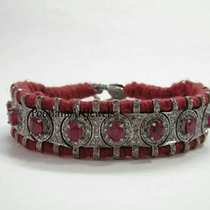 Può includere: Un braccialetto in corda rossa con una fascia argentata incastonata con rubini rossi rotondi e piccoli diamanti bianchi. La fascia è incisa con il testo "Poomam Art Jewels".