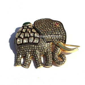 May include: A gold elephant brooch encrusted with small diamonds. The elephant has a green gemstone on its back and a red gemstone on its head. The text "PoonamArtJewels" is visible on the elephant's side.