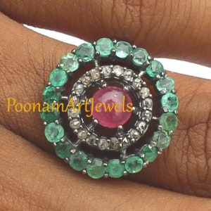 May include: A close-up of a ring featuring a central pink gemstone surrounded by a halo of small diamonds and a second halo of green gemstones. The ring is set on a dark metal band. The text "PoonamArtJewels" is visible.