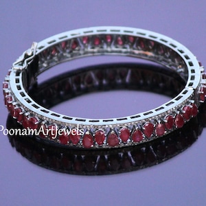 May include: A silver-toned hinged bangle bracelet with a row of teardrop-shaped red gemstones. The bracelet is set with smaller, clear stones and has a reflective surface.