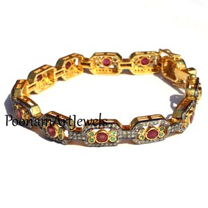Op de afbeelding: Een gouden en zilveren armband met een kettinglinkontwerp. De armband is versierd met rode robijnen en groene smaragden die in de schakels zijn gezet. De armband is geaccentueerd met kleine diamanten.