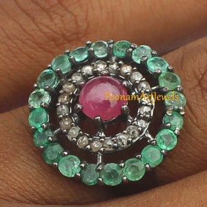 May include: A silver ring featuring a central, round, ruby gemstone surrounded by small diamonds and emeralds. The emeralds are arranged in a circular pattern around the ruby, creating a striking contrast of colors and textures. The ring is a piece of jewelry.