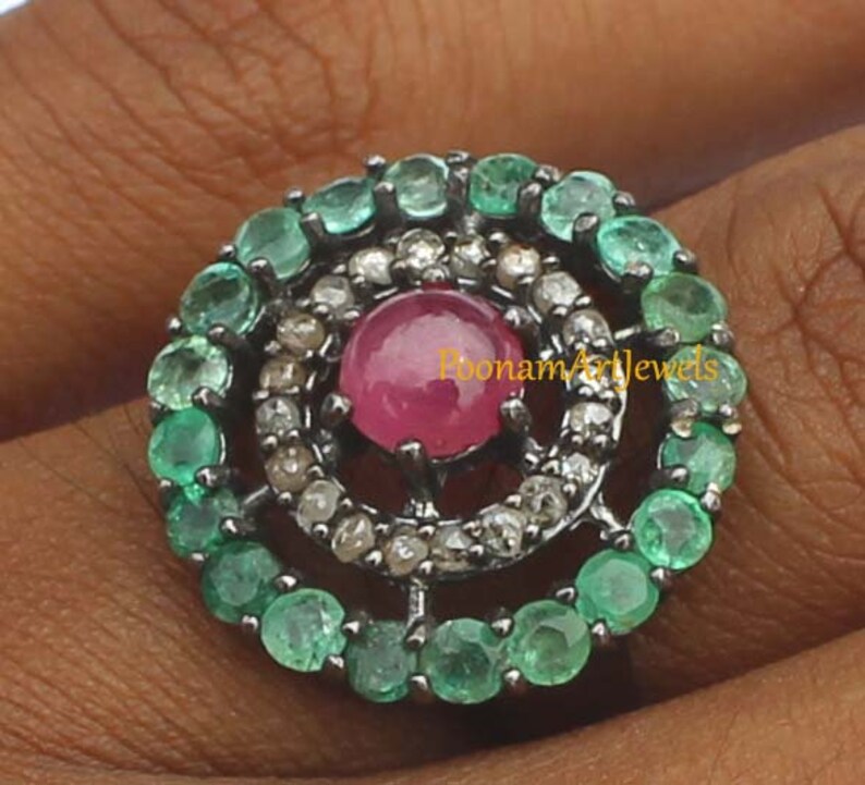 May include: A silver ring featuring a central, round, ruby gemstone surrounded by small diamonds and emeralds. The emeralds are arranged in a circular pattern around the ruby, creating a striking contrast of colors and textures. The ring is a piece of jewelry.
