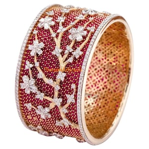 Puede incluir: Un brazalete de oro con un diseño floral engastado con piedras preciosas rojas y blancas. El brazalete está decorado con un delicado patrón de ramas y presenta una fila de pequeñas piedras blancas alrededor del borde.